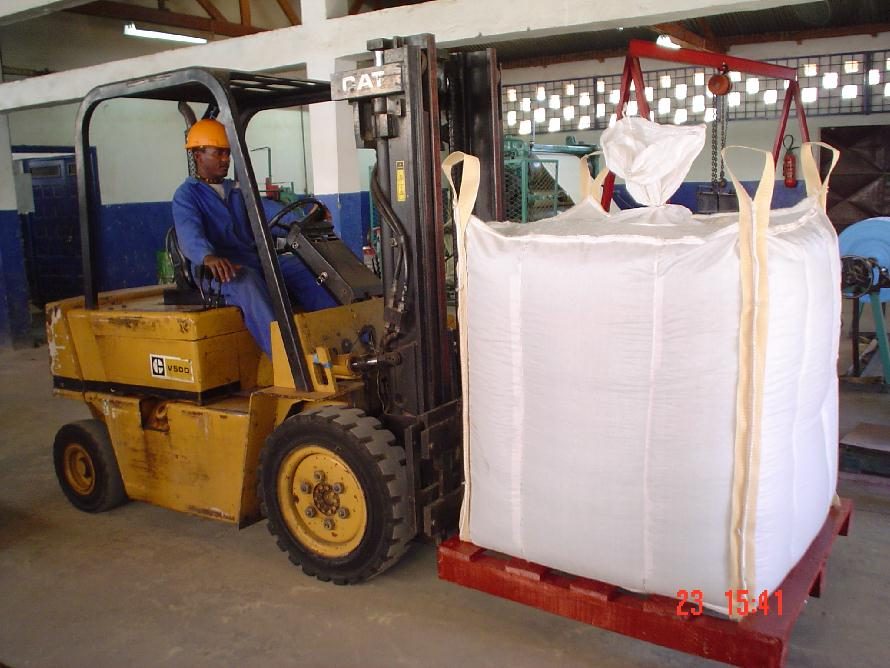 Forklift operator lifting a filled bulk bag with a spreader bar and chains in a warehouse